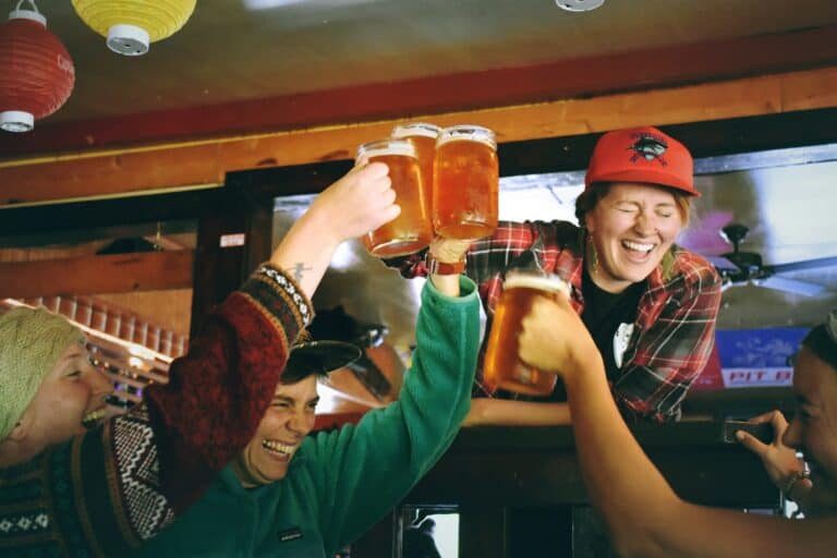 Four people raise their glasses of beer in a toast while smiling and laughing in a casual, warmly lit bar setting at Caliente Pizza & Drafthouse.