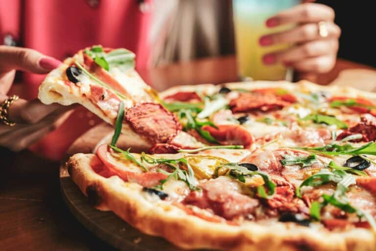 Person holding a slice of pizza with various toppings, including olives and arugula, from Pittsburgh Pizzeria, with a drink in the other hand.