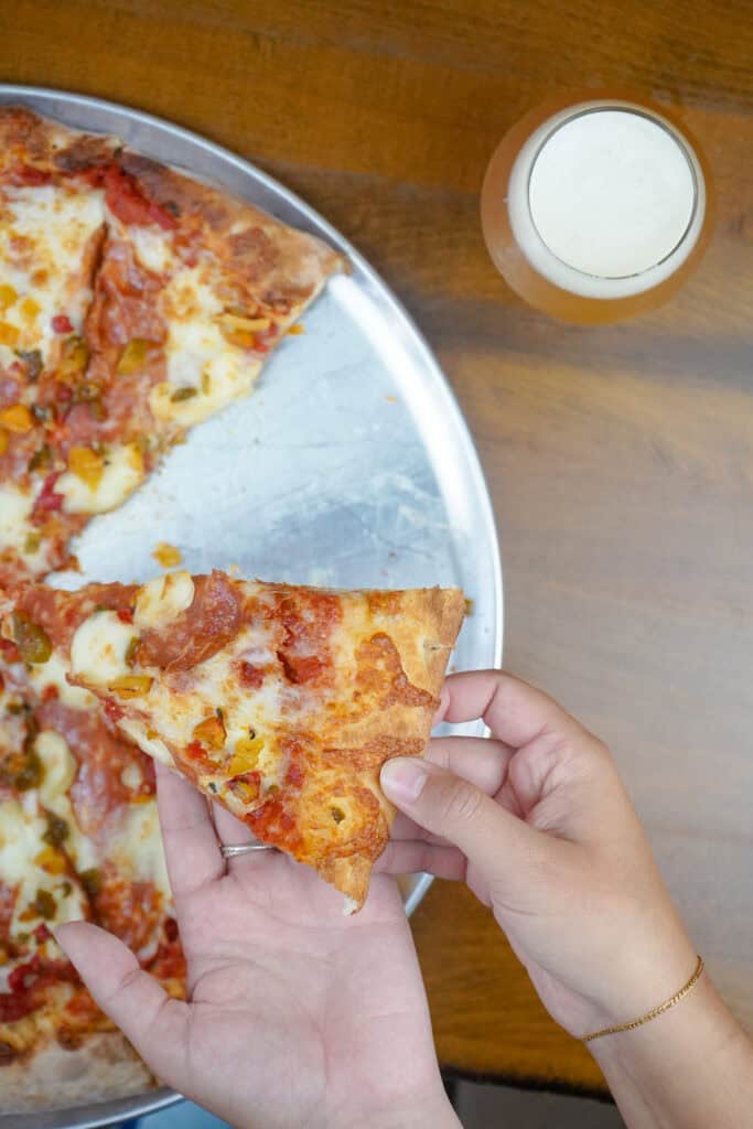 A person holds a slice of pizza above a metal tray with remaining slices. A glass of beer is nearby on a wooden table.
